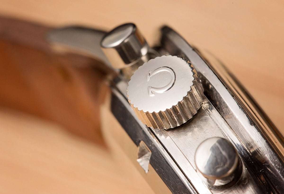 A close-up view of the crown and pushers on an Omega Speedmaster watch, showcasing the fluted design of the crown and the polished finish of the case, set against a warm, blurred background.