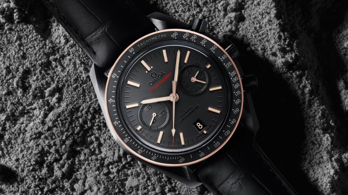 A close-up view of an Omega Speedmaster Moonwatch, featuring a black dial with rose gold-tone hands and indices, a tachymeter bezel, and a black leather strap, set against a textured gray background.