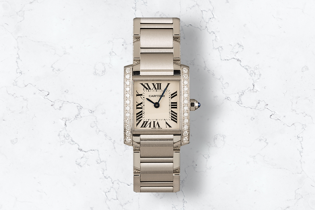 The image shows a silver-colored Cartier watch against a white marble background. The rectangular watch face features Roman numerals and is adorned with diamonds on the sides. The watch has a linked metal bracelet-style band