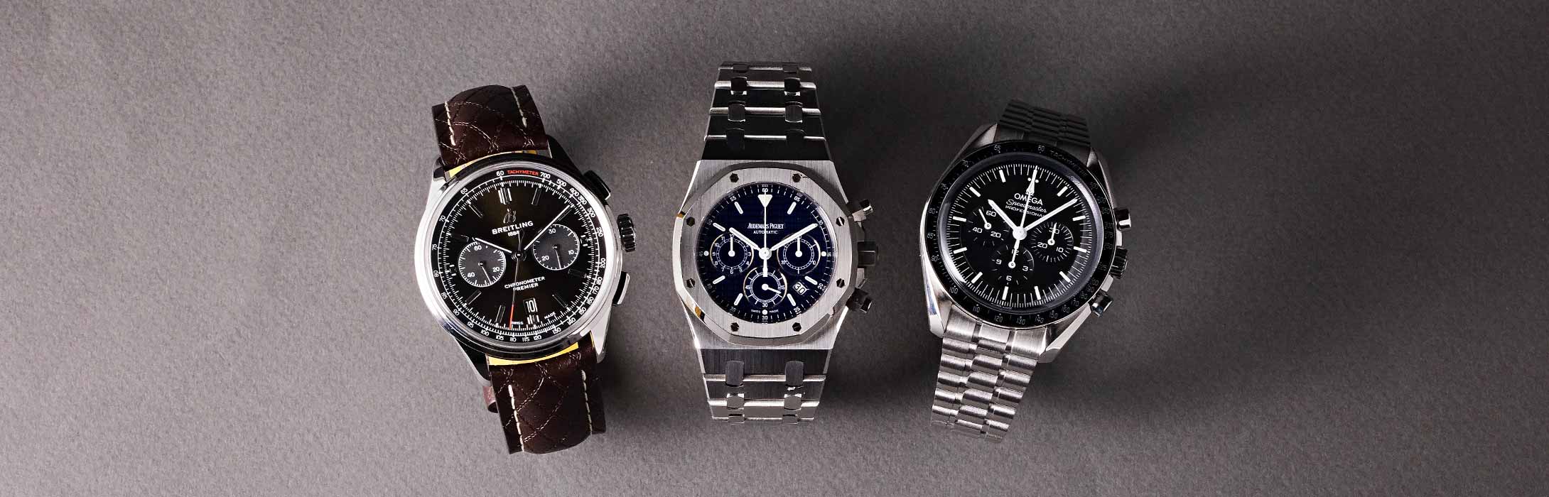 Three luxury watches - one black and gold chronograph, one stainless steel chronograph, and one stainless steel watch with black dial - displayed side-by-side on a gray background