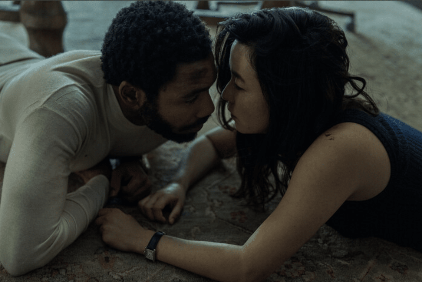 A romantic scene from Amazon's TV show 'Mr. and Mrs. Smith'. Actors lie close together on the floor, their faces nearly touching in an intimate moment. The man wears a white shirt, and the woman wears a dark sleeveless top.