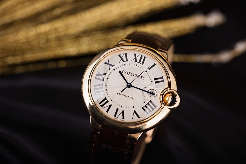 Cartier Ballon Bleu watch with a round gold case, silver dial featuring Roman numerals, blue sword-shaped hands, and a brown leather strap, elegantly displayed against a black and gold blurred background.