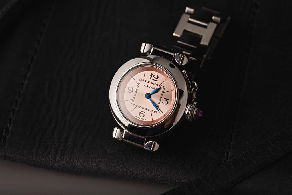 Cartier Pasha watch with a round stainless steel case, pink dial featuring Arabic numerals and blue sword-shaped hands, and a polished steel bracelet, displayed against a black leather background.