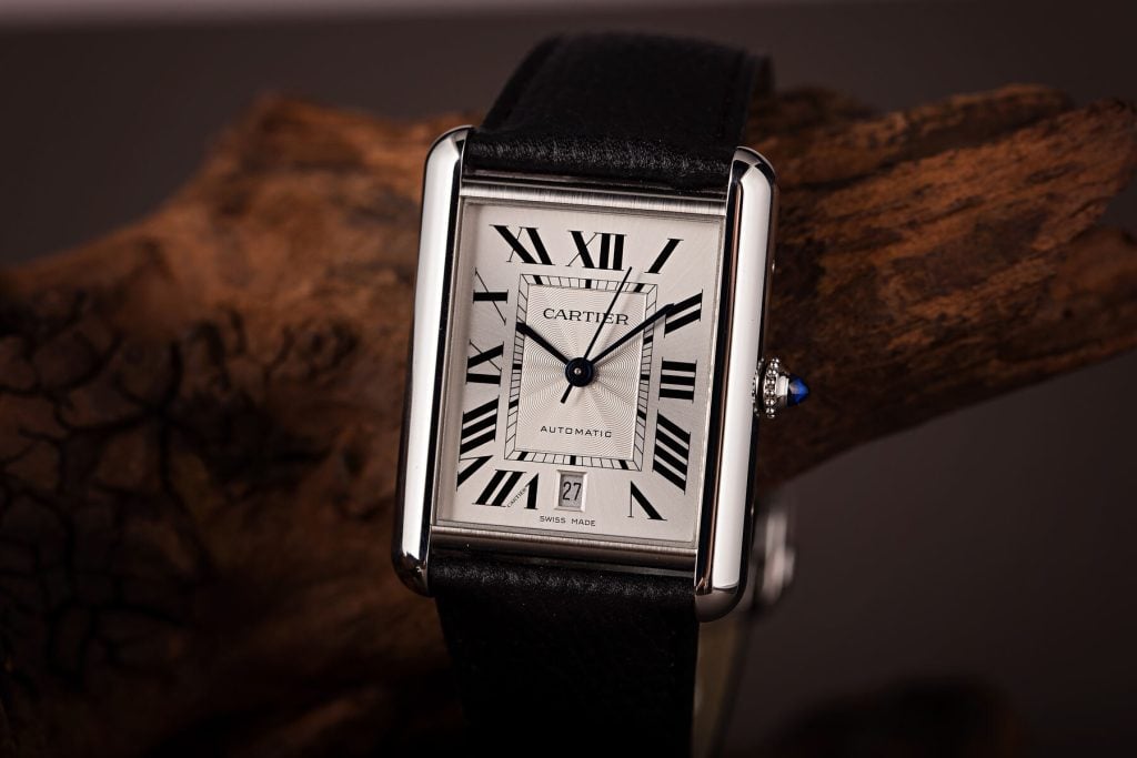 Cartier Tank watch with a rectangular silver dial, Roman numerals, black leather strap, and blue sapphire crown, elegantly displayed on a wooden backdrop.